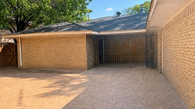 Brick paved front yard area with a metal gate enclosure and wooden side gate under a shingled roof with trees in background.