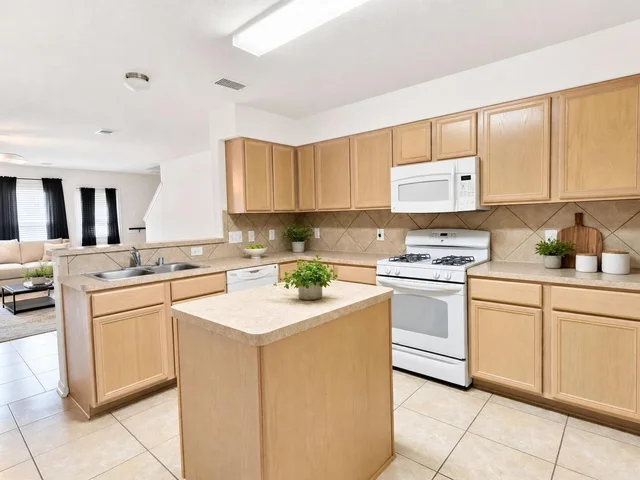 Kitchen with light wood cabinets, beige countertops, a kitchen island, white stove, microwave, and dishwasher under fluorescent light.