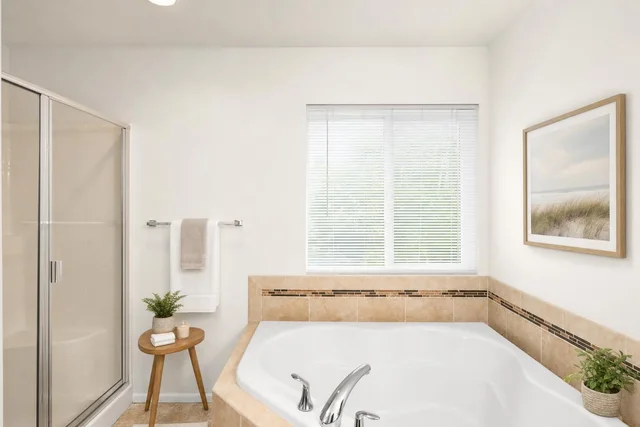 Bathroom with corner bathtub surrounded by beige tiles, glass shower enclosure, small wooden stool with towel and plant, and window with blinds.