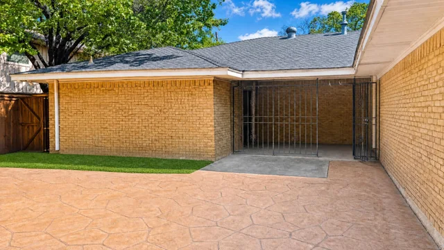 Stamped concrete driveway with yellow brick walls, an iron gate, a small patch of grass, and trees in the background under a blue sky.