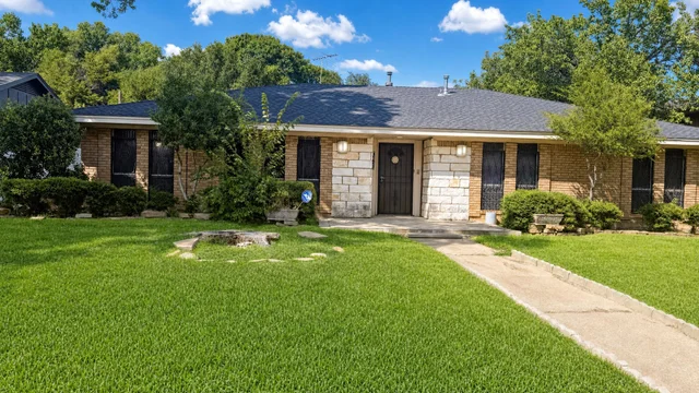 Single-story brick house with a black roof, concrete path, green grass lawn and bushes in the front yard on a sunny day.