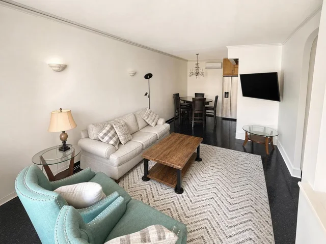 Modern living room with beige sofa, teal armchair, wooden coffee table on patterned rug, wall-mounted TV, dining table in background.