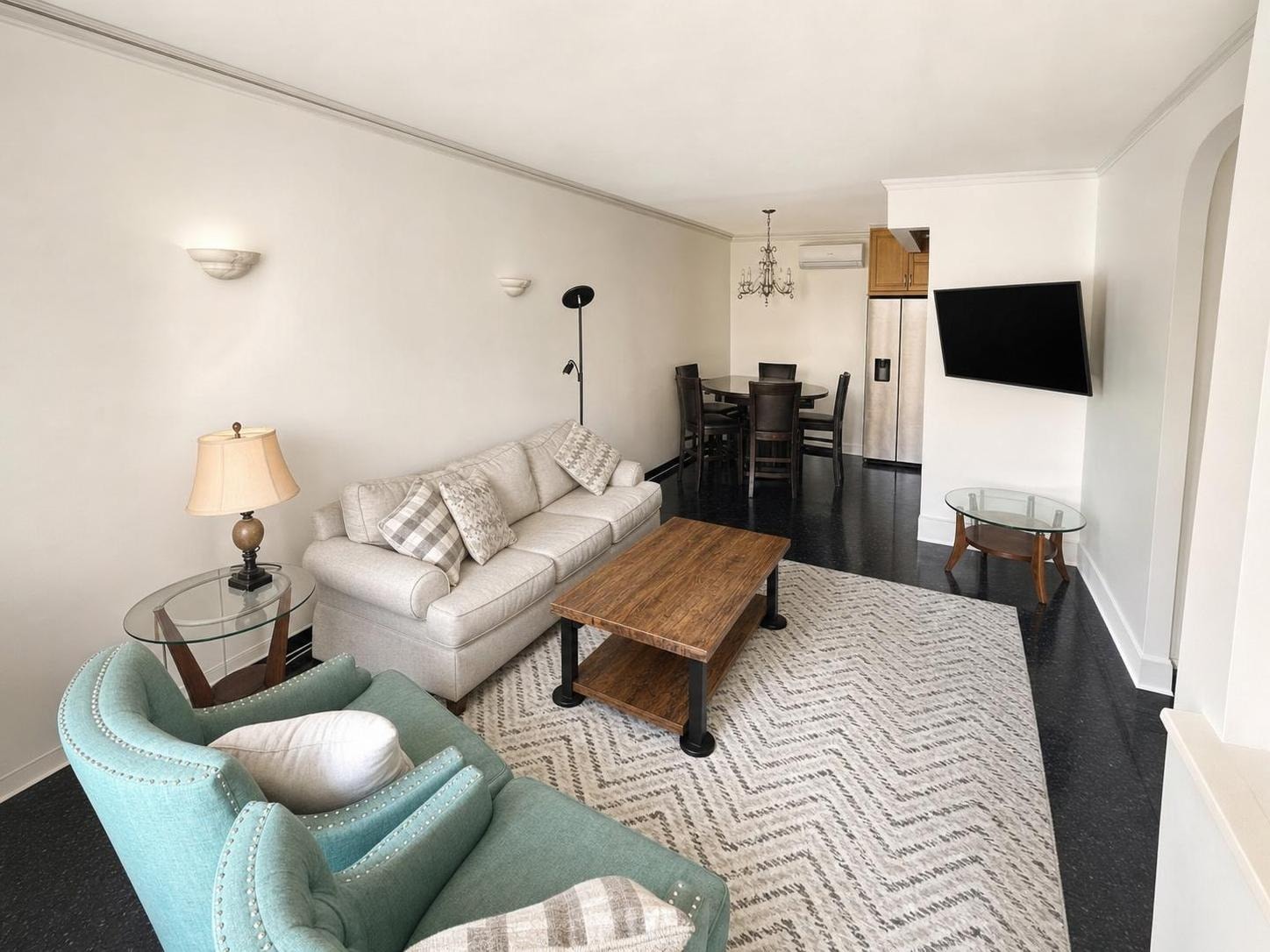 Modern living room with beige sofa, teal armchair, wooden coffee table on patterned rug, wall-mounted TV, dining table in background.