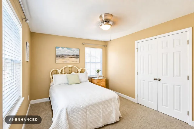 Bedroom with beige walls, white bed, wooden nightstand by window, and ceiling fan with light.