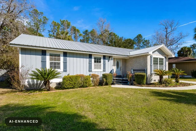Single-story house with white siding, gray metal roof, blue shutters, and a green front lawn with bushes.