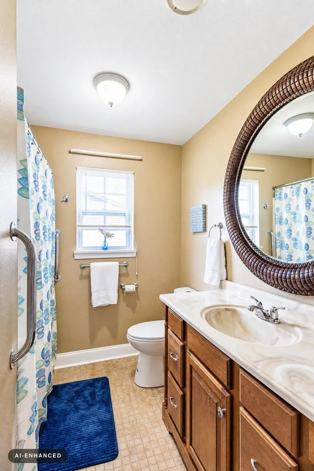 Small bathroom with wooden vanity, round mirror, toilet, blue bath mat, and blue patterned shower curtain.
