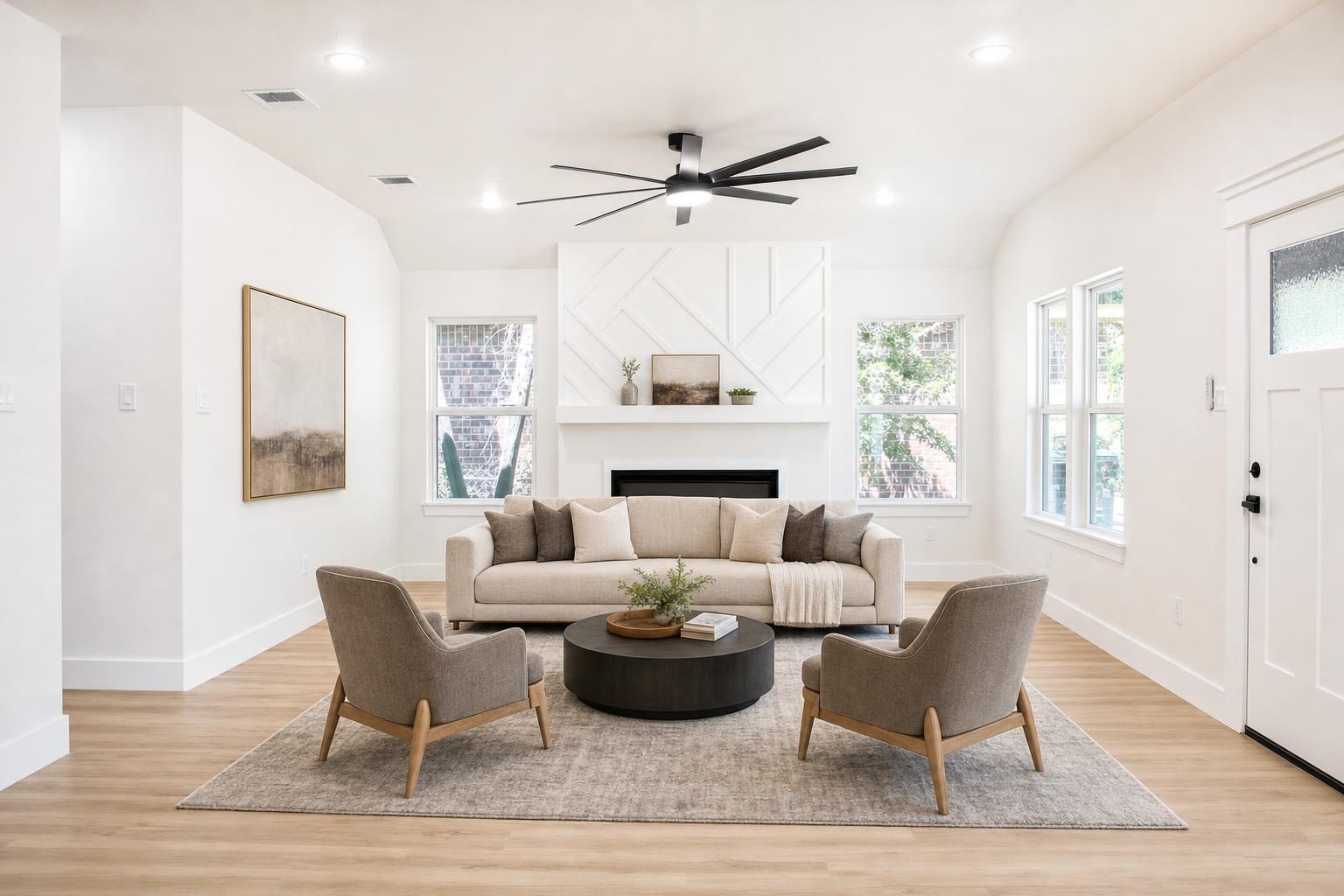 Bright modern living room with beige sofa, two gray armchairs, black round coffee table, light rug, white walls, and ceiling fan.