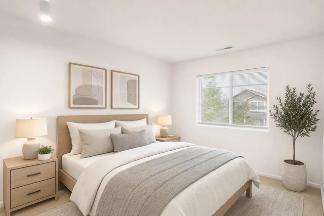 Bright bedroom with light wood bed, nightstands, two lamps, neutral bedding, abstract art above bed, large window, and a potted plant.