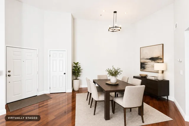 Modern dining room with dark wood table, six beige upholstered chairs on beige rug, dark wood sideboard with lamp, plant, and abstract wall art.
