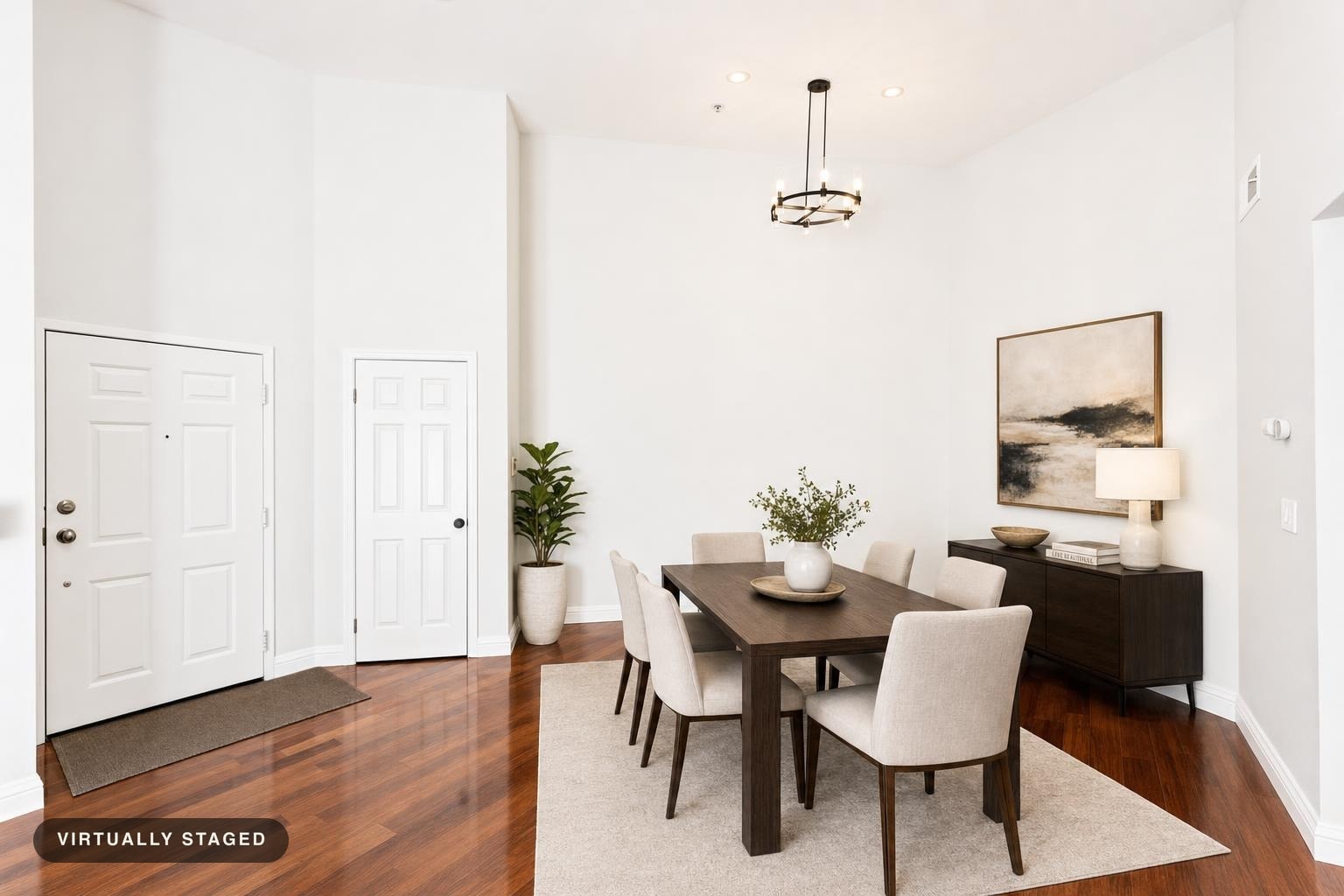 Modern dining room with dark wood table, six beige upholstered chairs on beige rug, dark wood sideboard with lamp, plant, and abstract wall art.