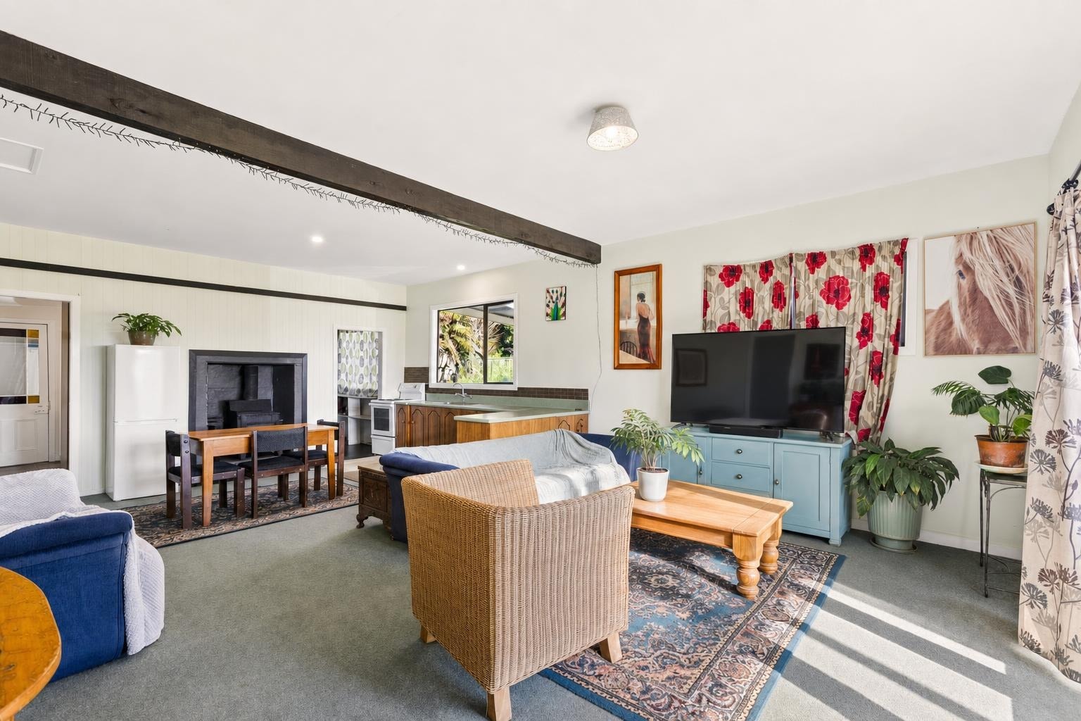 Living room with blue sofa, rattan chair, wooden coffee table, TV on a blue cabinet, and floral patterned curtains with sunlight.