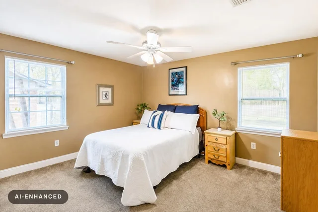Bedroom with tan walls, two windows, white bed linens with blue pillows, wooden nightstands, and a ceiling fan.