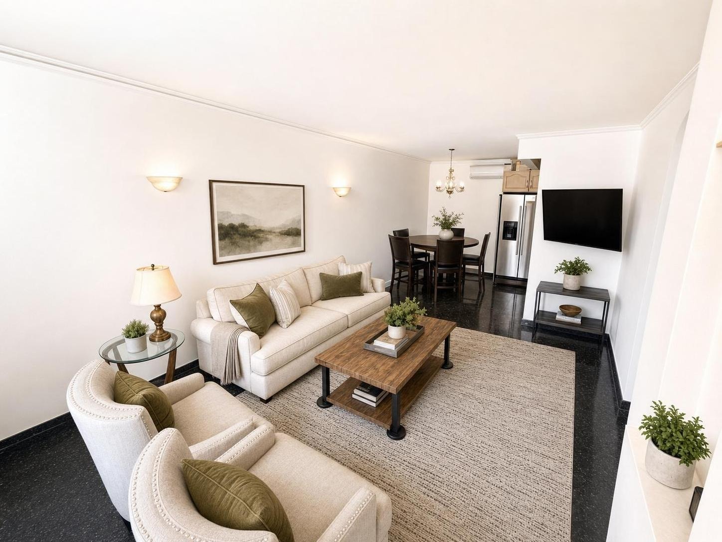 A modern living room with a beige sofa and armchair, wood and metal coffee table, wall-mounted TV, and dining table in the background.