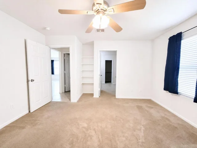 Empty bedroom with beige carpet, white walls, open doors, ceiling fan with lights, and blue curtains on the window.