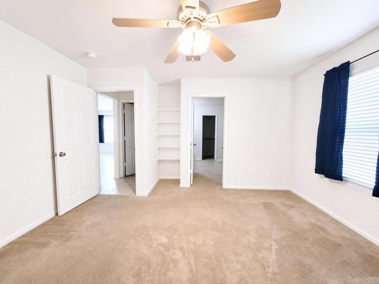 Empty bedroom with beige carpet, white walls, open doors, ceiling fan with lights, and blue curtains on the window.