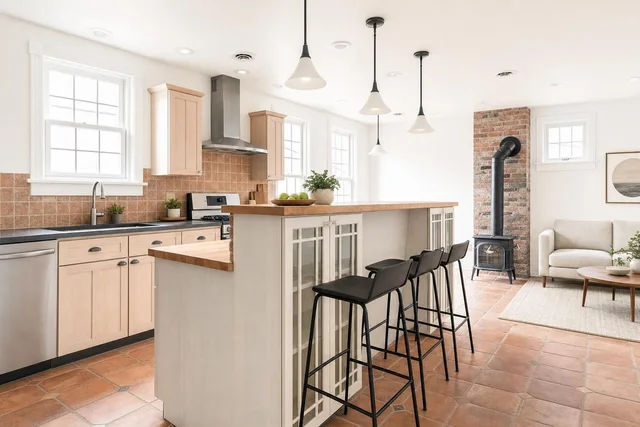 Bright modern kitchen with light wood cabinets, black stools at island counter, terracotta tile floor, and wood stove on brick wall.
