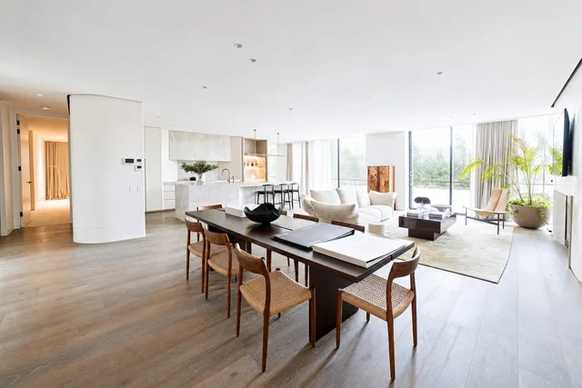 Bright modern living room with wood floor, white sofa, wooden dining table with chairs, large windows, and indoor plants.