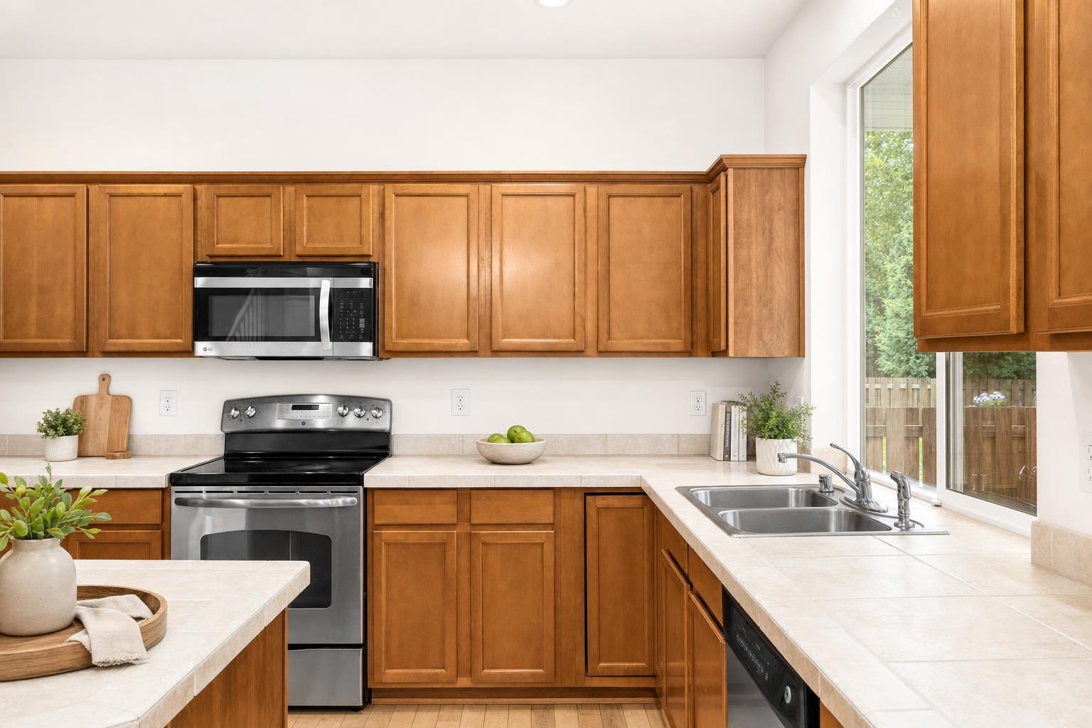 Kitchen with warm wood cabinets, stainless steel stove and microwave, beige tile countertops, and a large window over the sink.