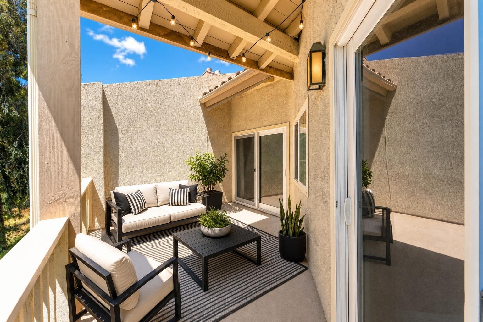 Modern backyard patio with black metal-framed sofa and chair with beige cushions, a striped rug, black coffee table, and potted plants.