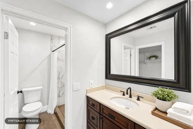 Modern bathroom showing dark wood vanity with beige countertop, large dark framed mirror, white toilet, and shower with white curtain.