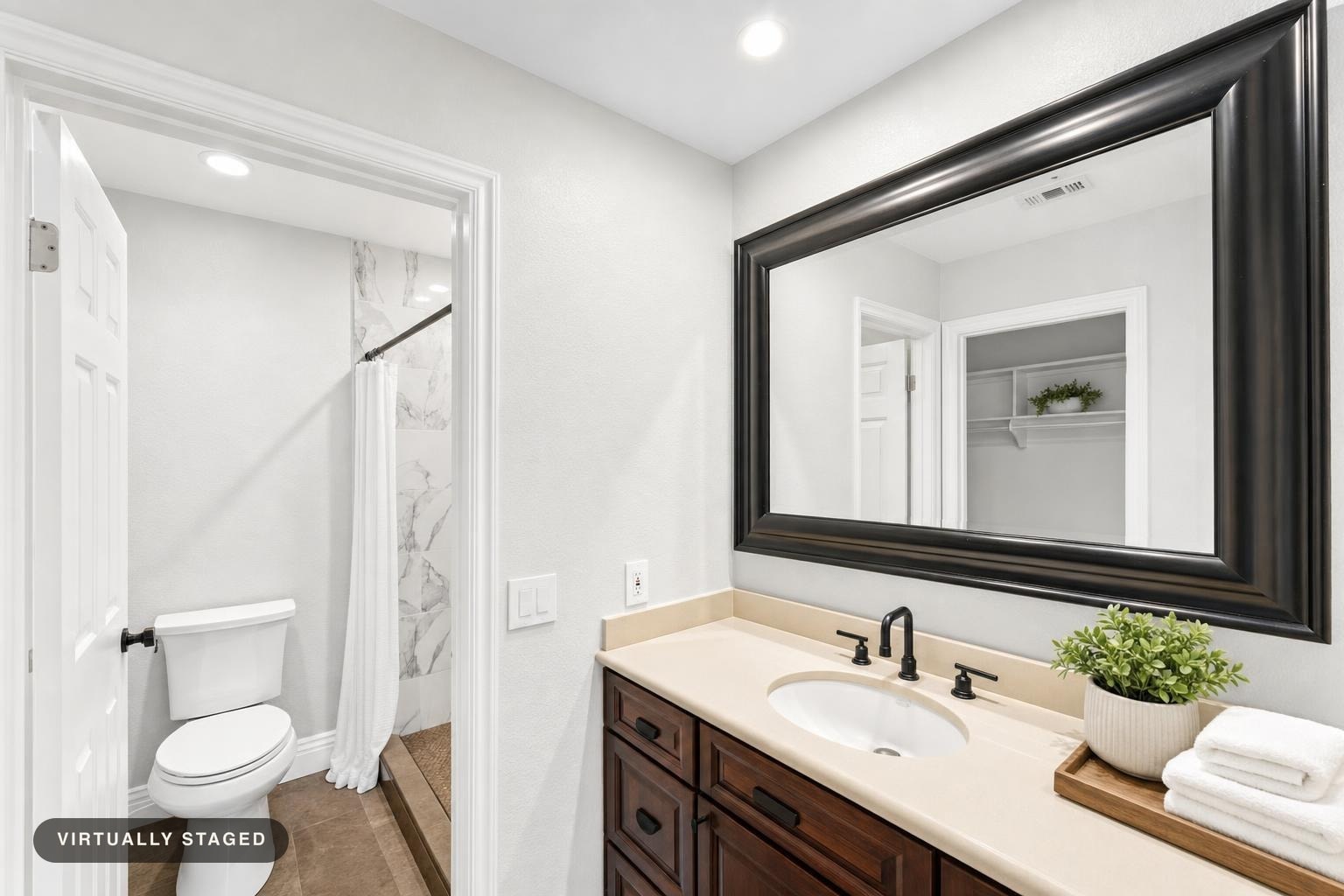 Modern bathroom showing dark wood vanity with beige countertop, large dark framed mirror, white toilet, and shower with white curtain.