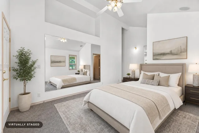 Modern bedroom with beige upholstered bed, large wall mirror, potted plant, two nightstands with lamps, and a ceiling fan.