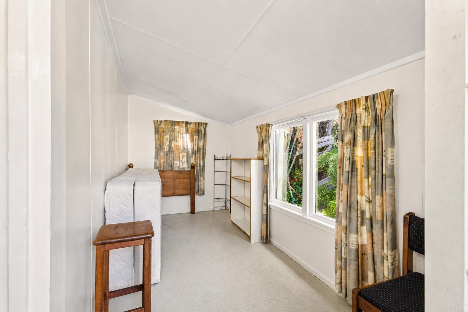 Small bedroom with two windows, floral patterned curtains, mattress leaning against wall, wooden chair and shelves, white walls.
