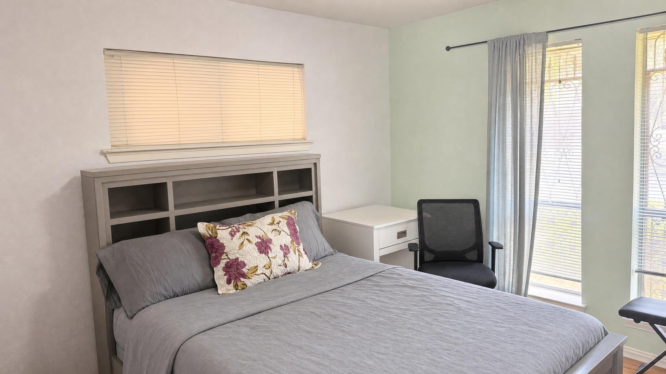 Modern bedroom with gray bed, floral pillow, white nightstand, black office chair by window with sheer curtains.