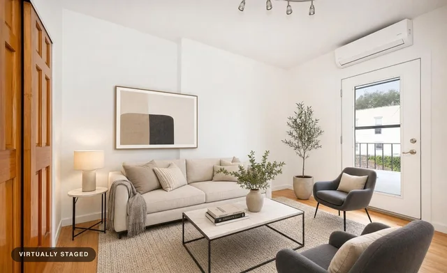 Modern living room with beige sofa, two black accent chairs, a light rug, two potted plants, a side table with lamp, and abstract wall art.