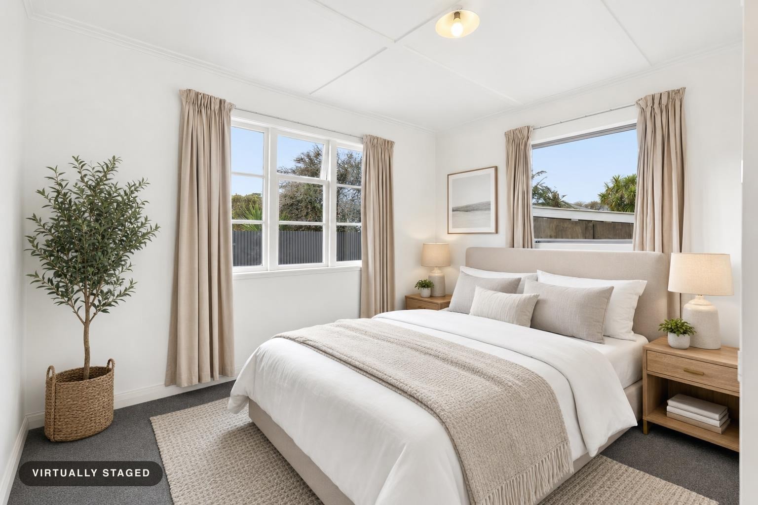 Bright bedroom with beige upholstered bed, wooden nightstands with lamps, potted plant, and beige curtains on windows.