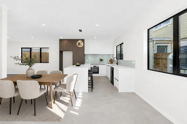Bright modern dining area with wooden rectangular dining table, beige upholstered chairs, and adjacent white kitchen with wood accent cabinet.