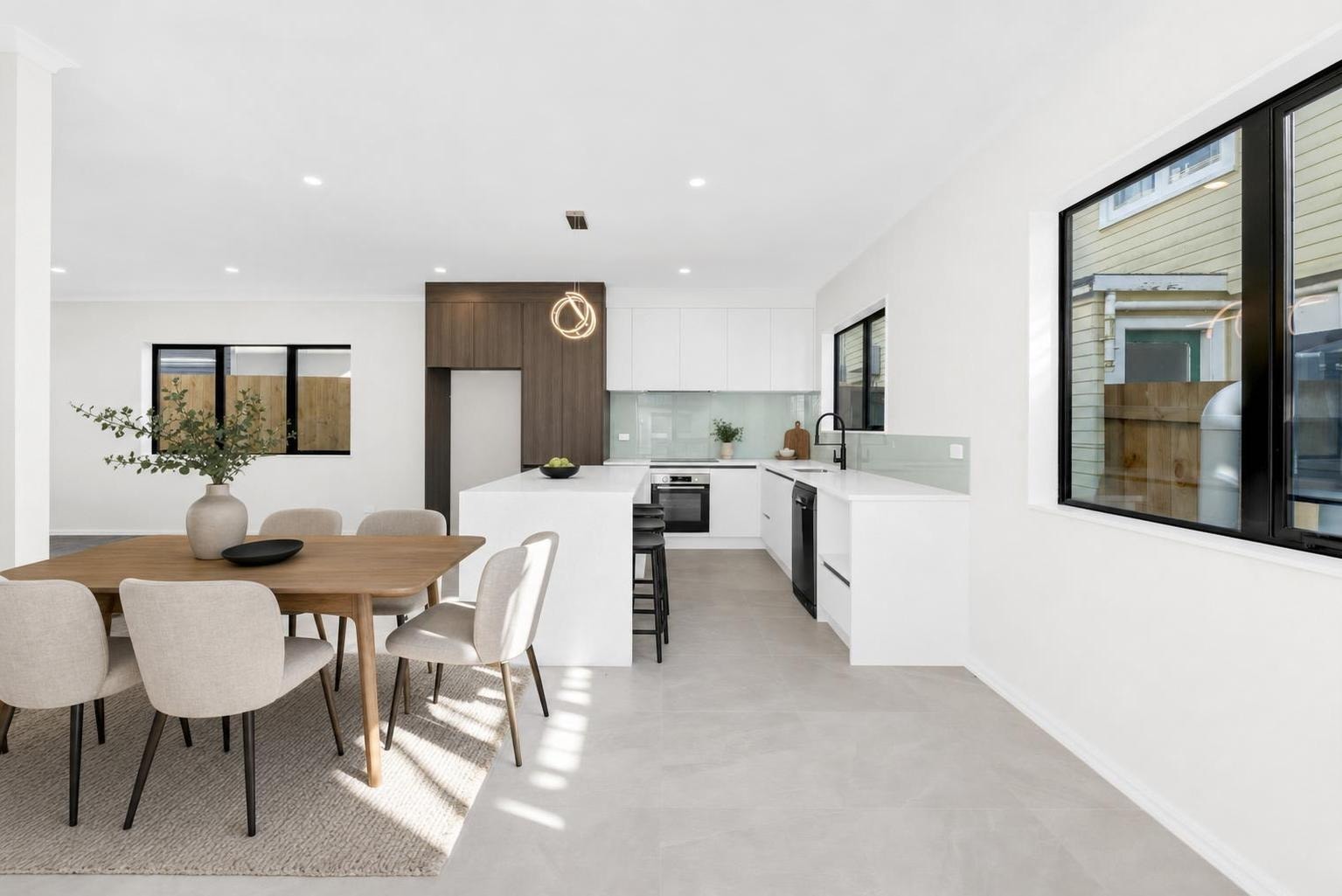 Bright modern dining area with wooden rectangular dining table, beige upholstered chairs, and adjacent white kitchen with wood accent cabinet.
