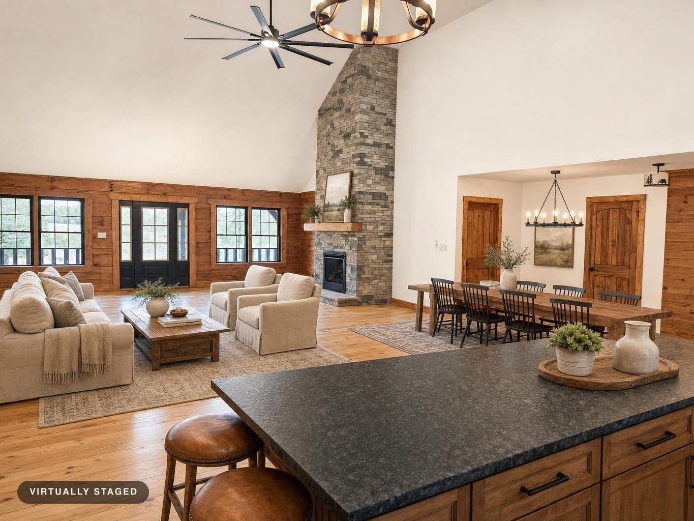 Spacious living room with beige sofa and armchairs, wooden coffee and dining tables, stone fireplace, and black ceiling fan.