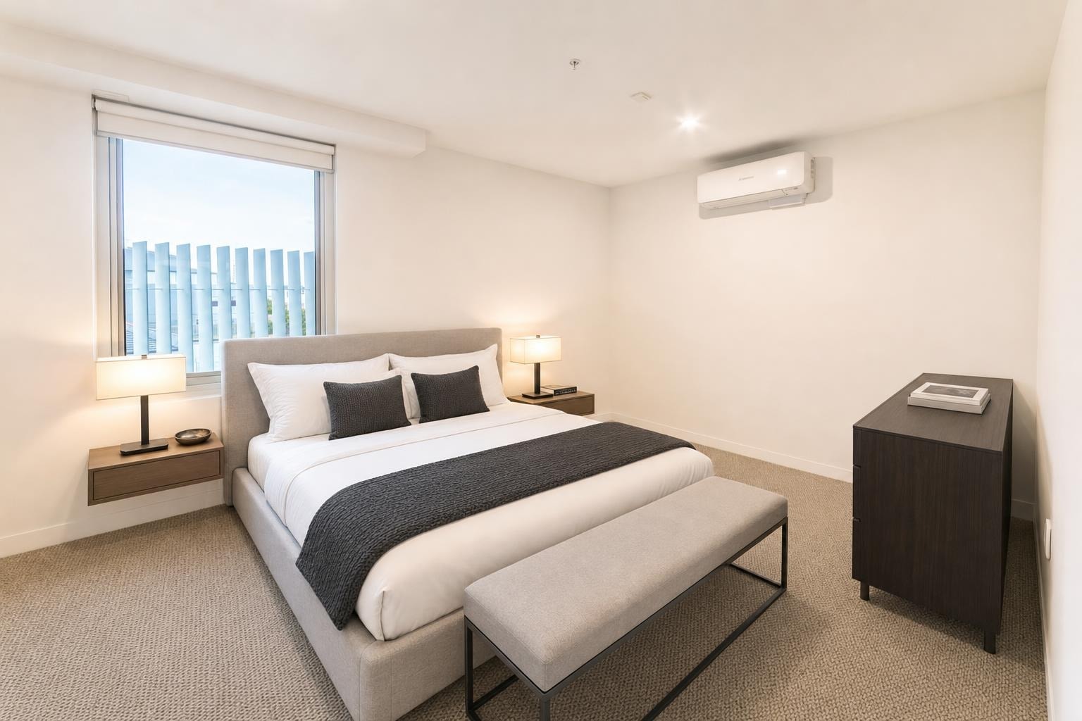 Modern bedroom with gray bed, gray bench, floating nightstands with lamps, dark dresser, and window with vertical blinds.