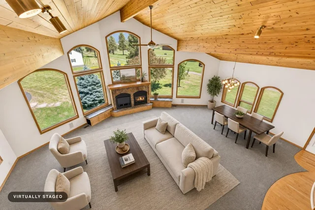Living room with vaulted wood ceiling, large arched windows, beige sofa, two armchairs, wooden coffee table, dining table with six chairs, and a stone fireplace.