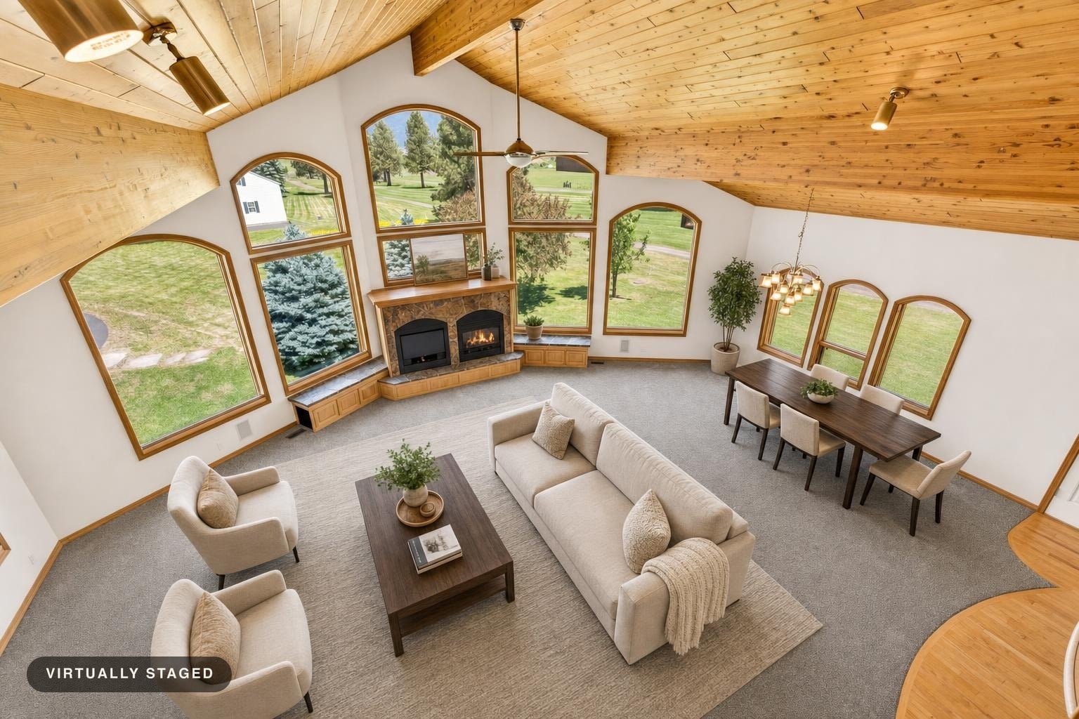 Living room with vaulted wood ceiling, large arched windows, beige sofa, two armchairs, wooden coffee table, dining table with six chairs, and a stone fireplace.