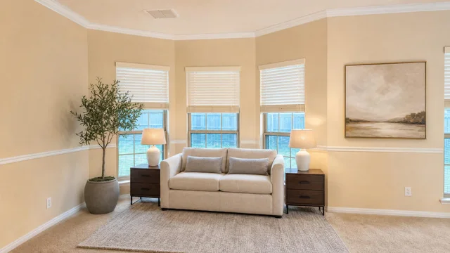 Living room with beige loveseat between two dark wood nightstands with lamps, large plant on the left, and a neutral painting on the wall.