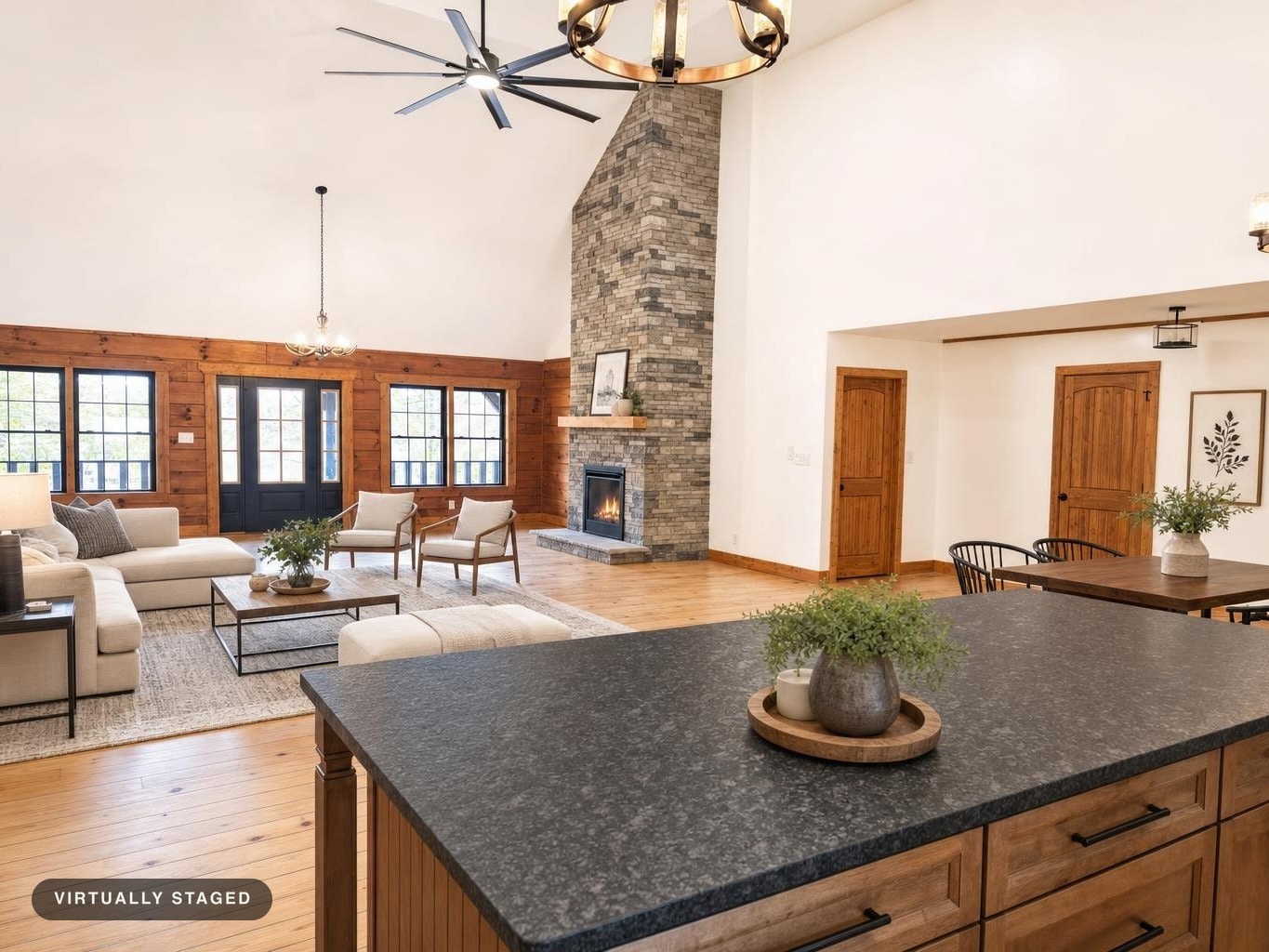 Living room with beige sectional sofas, stone fireplace, wooden doors and trims, black metal chandelier, and large black countertop in foreground.