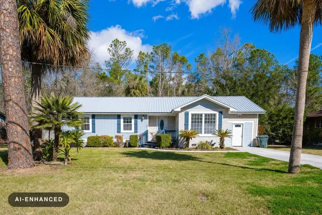 Front view of a single-story house with metal roof, blue shutters, small porch, palm trees, and green lawn under a blue sky.