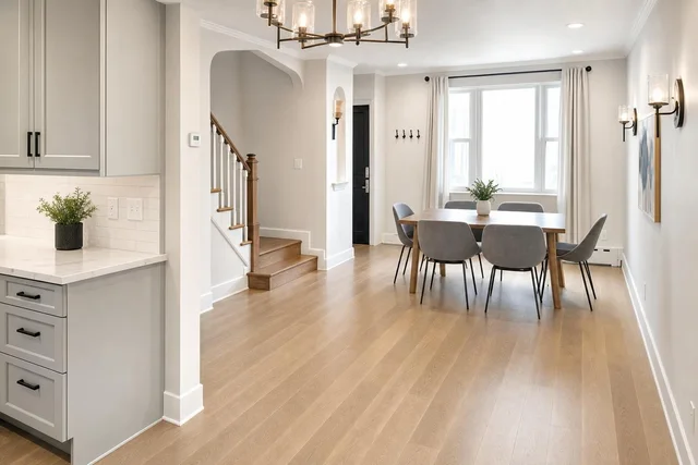 Dining room with wooden table, six grey chairs with black metal legs, wood floor, white walls, and natural light from window.