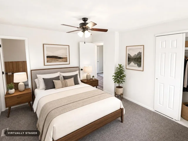 Modern bedroom with a wooden bed, neutral bedding, matching nightstands with lamps, framed landscape prints, and a ceiling fan.