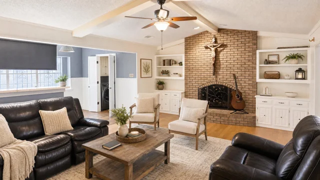 Farmhouse living room with brown leather sofas, wooden coffee table, two armchairs, brick fireplace, and ceiling fan.
