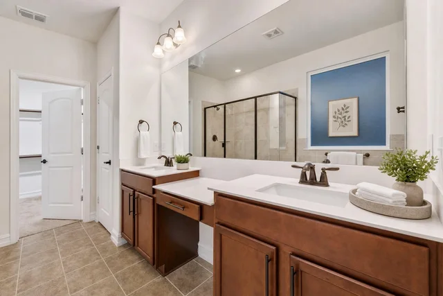 Modern bathroom with double white sinks on brown wood cabinets, large mirror, glass shower with black frame, and blue accent wall artwork.