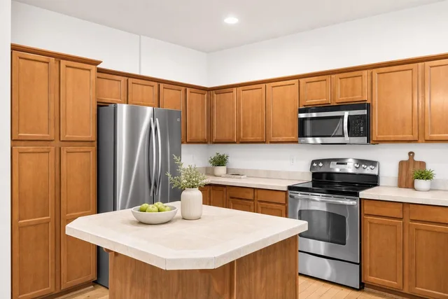 Kitchen with brown wood cabinets, stainless steel refrigerator, stove, microwave, and central island with vase and bowl of green apples.