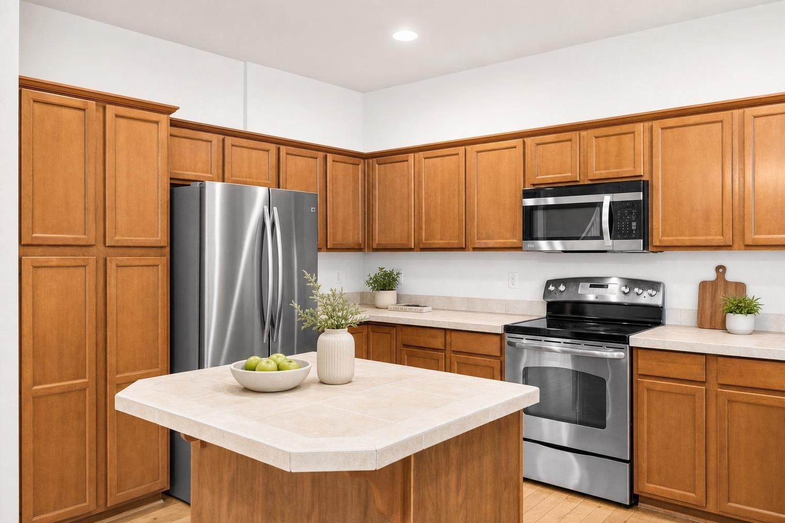 Kitchen with brown wood cabinets, stainless steel refrigerator, stove, microwave, and central island with vase and bowl of green apples.