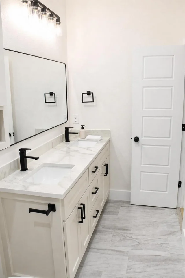 White bathroom with a marble-look double sink countertop, black faucets, black cabinet handles, and a white door.