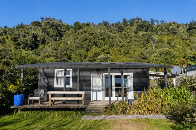 Black wooden house with white-framed windows and door, wooden porch with bench and chair, green plants and grassy yard in front