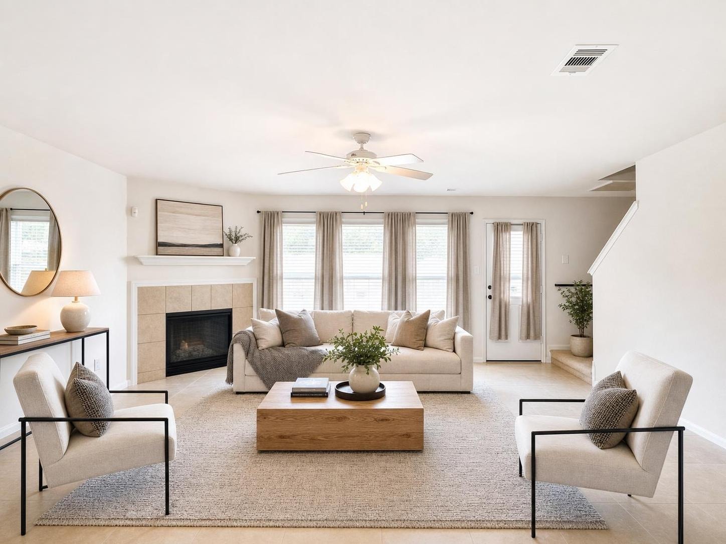 Bright modern living room with beige sofa, two armchairs, wooden coffee table, fireplace, and ceiling fan.