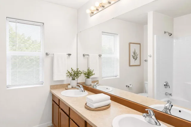 White bathroom with double sink vanity, tan countertop, two windows with blinds, white towels, and a framed leaf print.