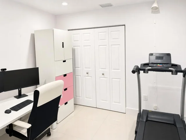 A modern office with a black and white chair, white desk with monitor, pink and white cabinet, and a treadmill against a white wall.
